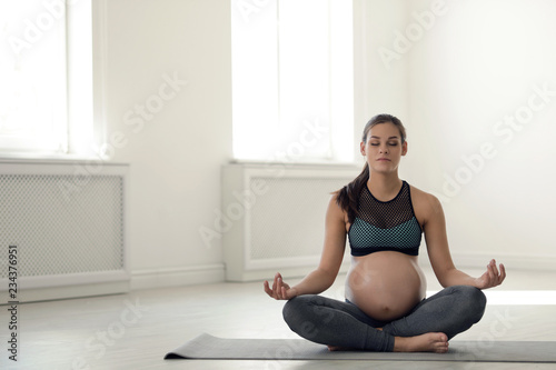Young pregnant woman in fitness clothes meditating at home. Space for text