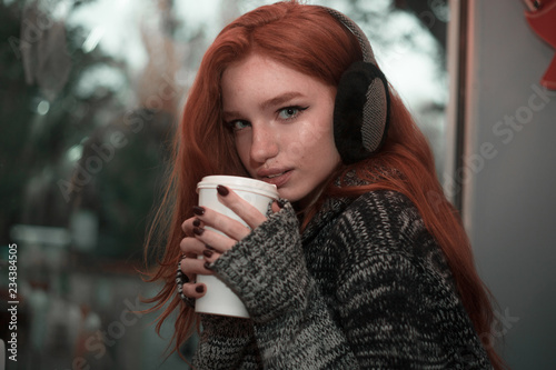 The portrait of a redhead smilling girl with cup of coffee in gray sveater sitting in a cafe pleased photo