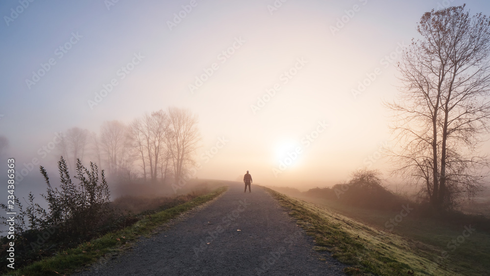 Man standing on a long gravel path with a foggy sunrise in the distance.
