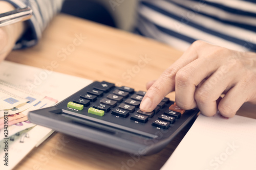 Cost calculation, payment day concept, hand put finger on calculator and other hand holding pen and pile of Euro banknotes with calendar on wood table, soft vintage effect