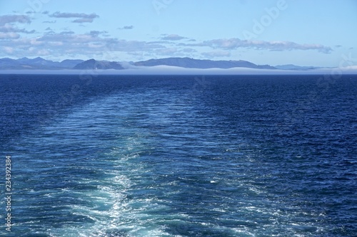 Morning at sea: Clouds and fog in a deep blue sky over Alaskan islands in the North Pacific Ocean, behind the wake of a cruise ship.