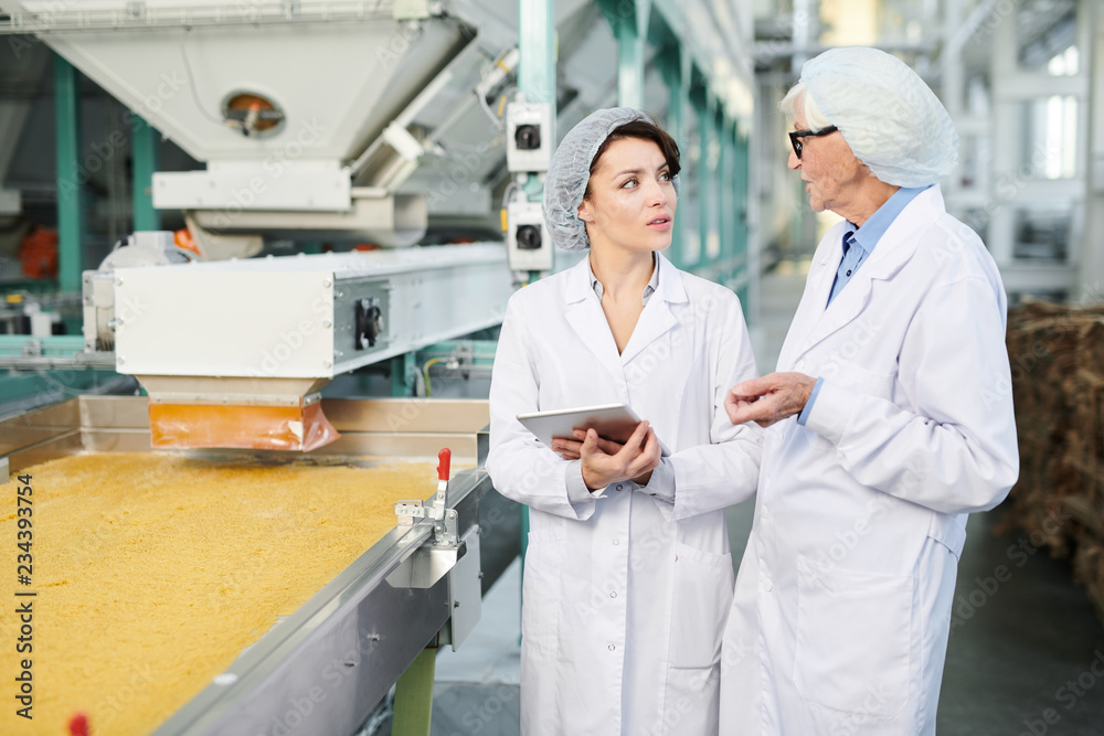 Portrait  of  two  female workers doing  production quality inspection in food factory standing by conveyor belt and using digital tablet, copy space
