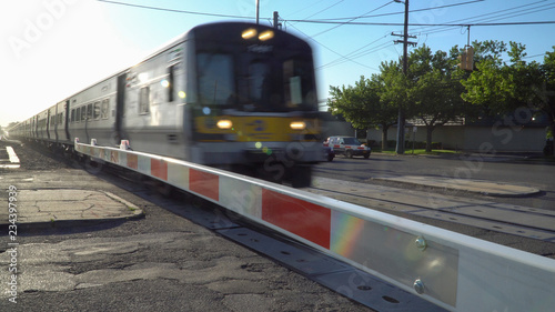 Railroad train crossing safety gate xing at local station. Motion blur high speed public transit commute. Morning sunrise lens flare.
