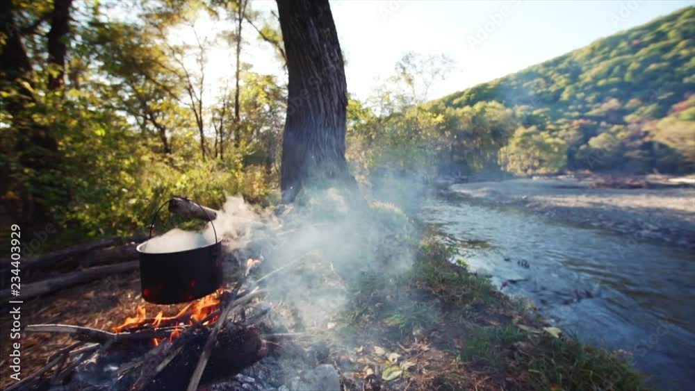 Peaceful camping under big tree: burning campfire and pot above it. Slow motion. Fast blue stream and autumn forest are on the background. Far East of Russia. Lovely sunny day Peaceful camping under big tree: burning campfire and pot above it. Slow motion. Fast blue stream and autumn forest are on the background. Far East of Russia. Lovely sunny day