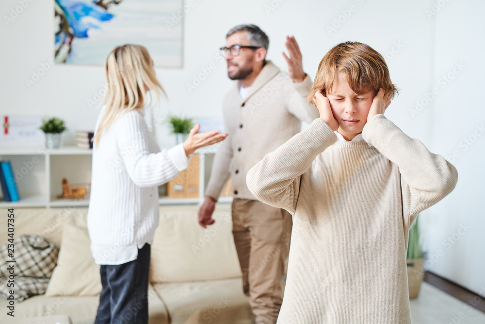 Fototapeta premium Distraught teenage son in sweater suffering from parents conflict: he keeping eyes closed and plugging in ears with fingers while his nervous parents fighting in background