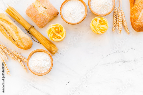 Farinaceous food. Fresh bread and raw pasta near flour in bowl and wheat ears on white stone background top view space for text photo