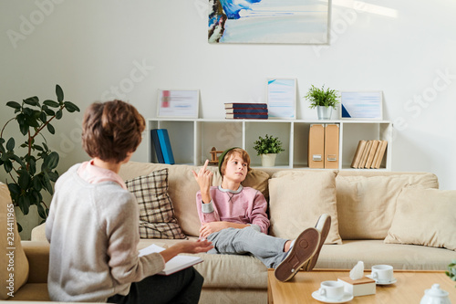 Photography Serious aggressive teenager in hat sitting on sofa and showing middle finger whi