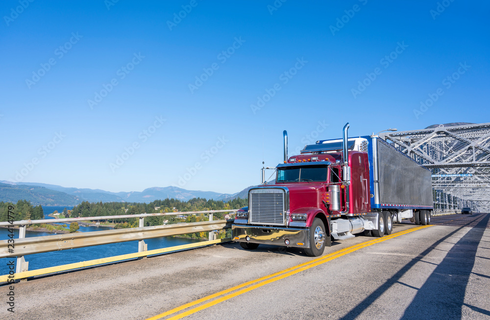 Burgundy big rig classic semi truck with refrigerated shiny semi trailer transporting commercial cargo on the Bridge of God in Columbia River Gorge