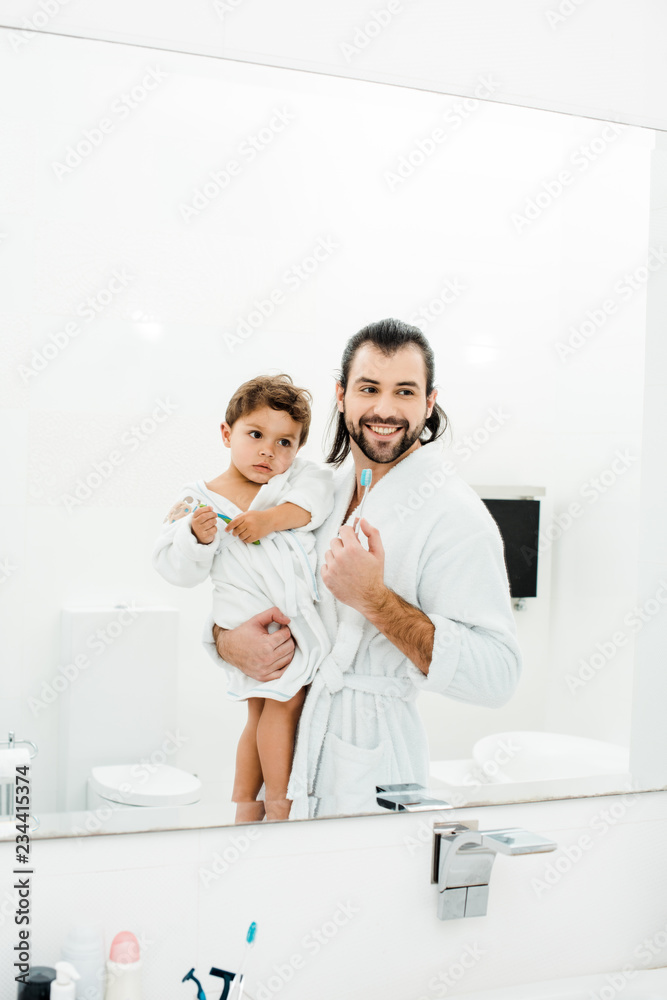 Fototapeta premium Dad and toddler son looking in mirror at home in white bathroom