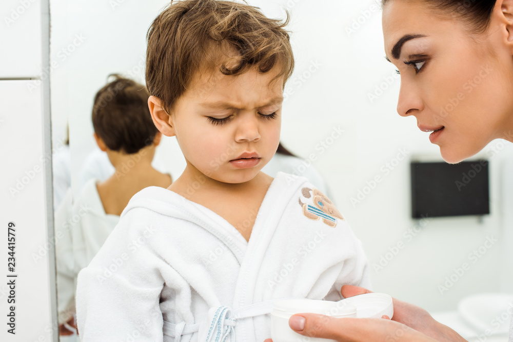mother holding face cream while son sitting in white bathrobe with closed eyes mother holding face cream while son sitting in white bathrobe with closed eyes