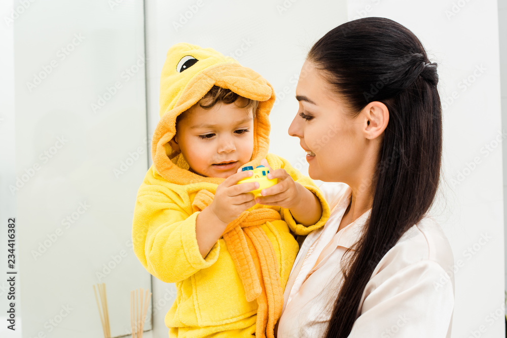 beautiful woman holding son in yellow bathrobe with rubber toy beautiful woman holding son in yellow bathrobe with rubber toy