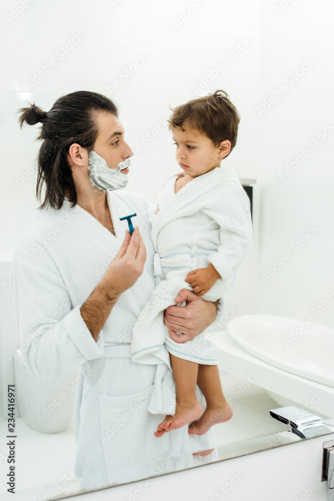 Dad in white bathrobe showing razor to toddler son Dad in white bathrobe showing razor to toddler son