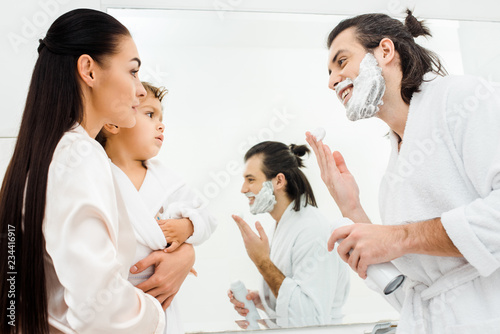 smiling man with foam on face looking at wife and son in bathroom