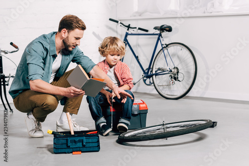 man with book pointing by hand at bicycle wheel to little son in workshop