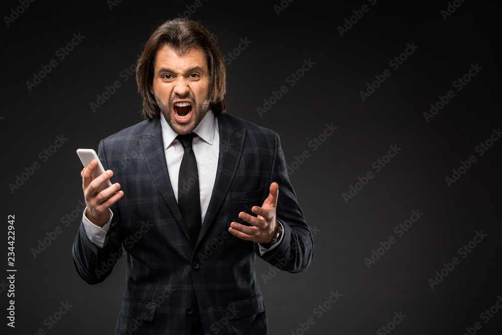 angry young businessman holding smartphone and yelling at camera isolated on black angry young businessman holding smartphone and yelling at camera isolated on black