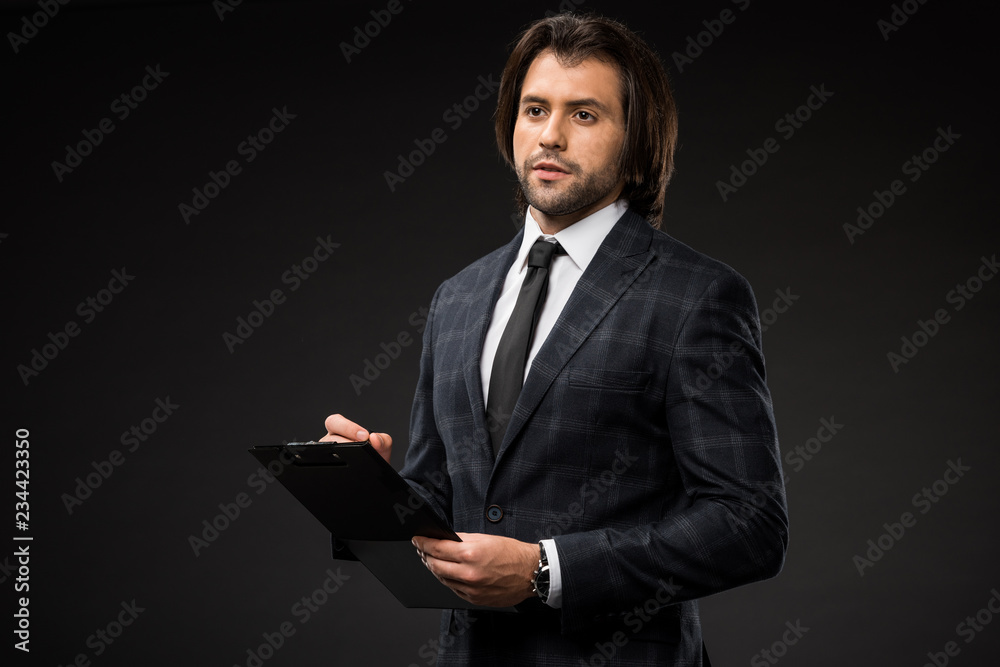 serious young businessman writing on clipboard and looking away isolated on black serious young businessman writing on clipboard and looking away isolated on black
