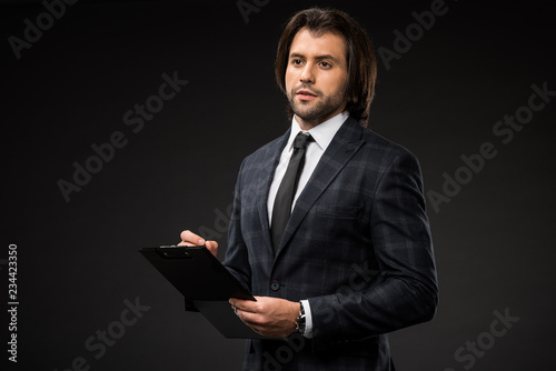 serious young businessman writing on clipboard and looking away isolated on black