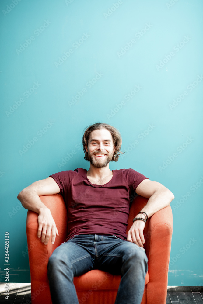 Portrait of a young caucasian bearded man with long hair dressed in t-shirt and jeans sitting on the chair on the colorful background. Image with copy space Portrait of a young caucasian bearded man with long hair dressed in t-shirt and jeans sitting on the chair on the colorful background. Image with copy space