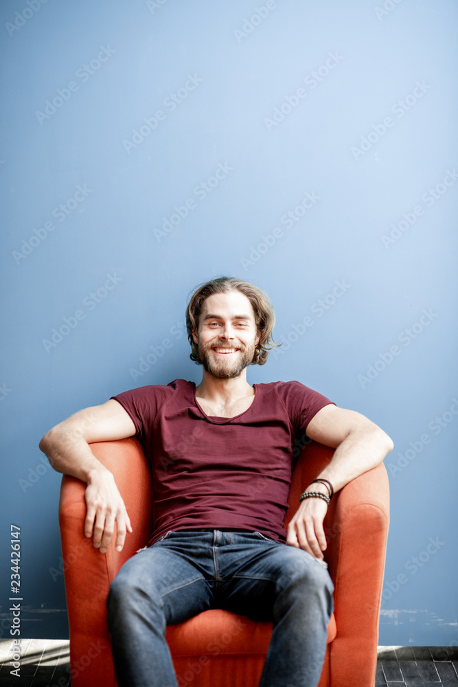 Portrait of a young caucasian bearded man with long hair dressed in t-shirt and jeans sitting on the chair on the colorful background. Image with copy space