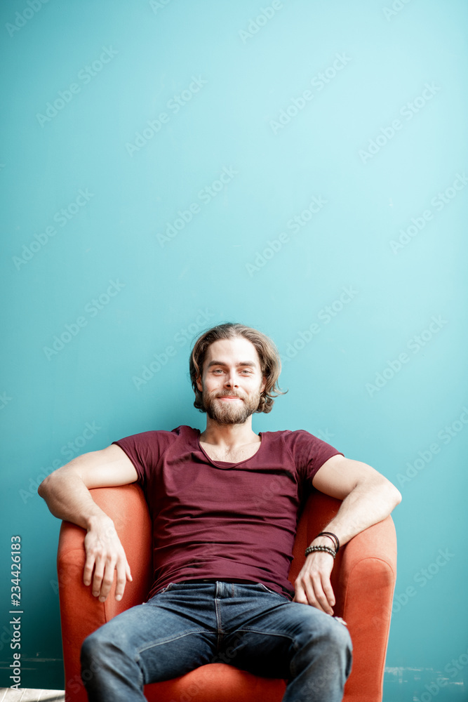 Portrait of a young caucasian bearded man with long hair dressed in t-shirt and jeans sitting on the chair on the colorful background. Image with copy space Portrait of a young caucasian bearded man with long hair dressed in t-shirt and jeans sitting on the chair on the colorful background. Image with copy space