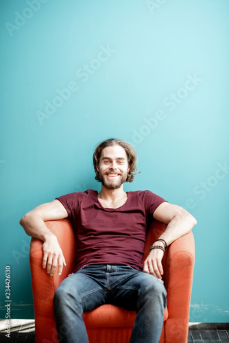 Portrait of a young caucasian bearded man with long hair dressed in t-shirt and jeans sitting on the chair on the colorful background. Image with copy space © rh2010