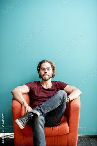 Portrait of a young caucasian bearded man with long hair dressed in t-shirt and jeans sitting on the chair on the colorful background. Image with copy space © rh2010