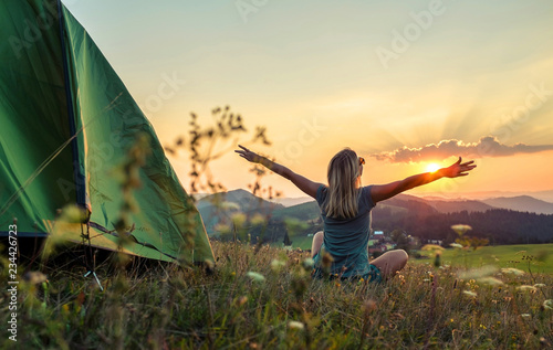 Happy woman with open arms stay near tent around mountains under sun