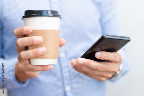 Closeup of business man holding smartphone and drink. Person standing and having break. Communication and break concept. Cropped front view. photo