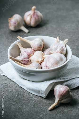 Garlic bulbs in ceramic bowl with white cloth on grey background