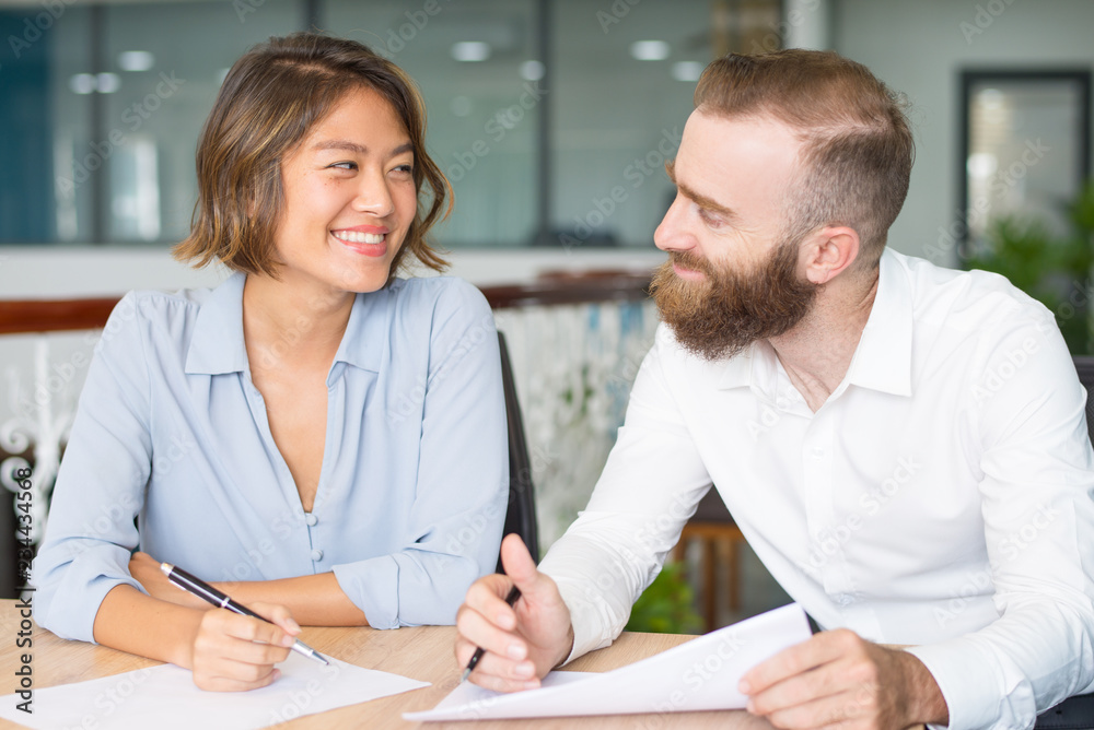 Positive analysts joking while checking reports. Two business colleagues discussing papers and smiling at each other. Teamwork concept Positive analysts joking while checking reports. Two business colleagues discussing papers and smiling at each other. Teamwork concept
