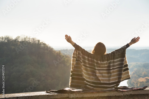 Young woman enjoying sunny day sitting on a hill. Raised hands. Back view