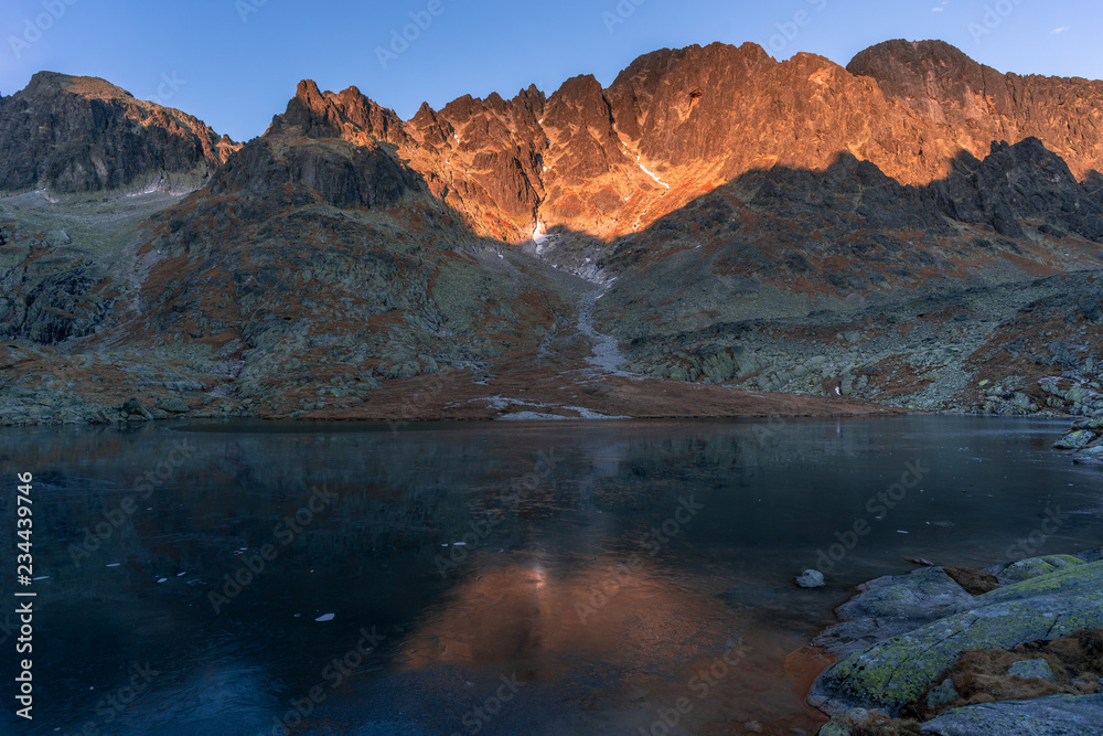 Last rays of sun setting fire to a high mountain ridge, with the frozen surface of the lake below