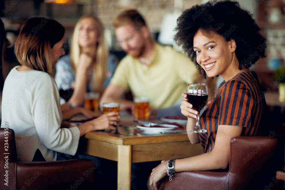 Woman looking at camera and holding glass of wine while sitting in restaurant. In background friends drinking and chatting.