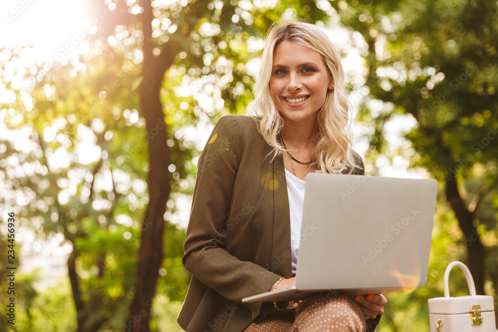 Image of attractive woman 20s using silver laptop, while sitting on bench in park Image of attractive woman 20s using silver laptop, while sitting on bench in park