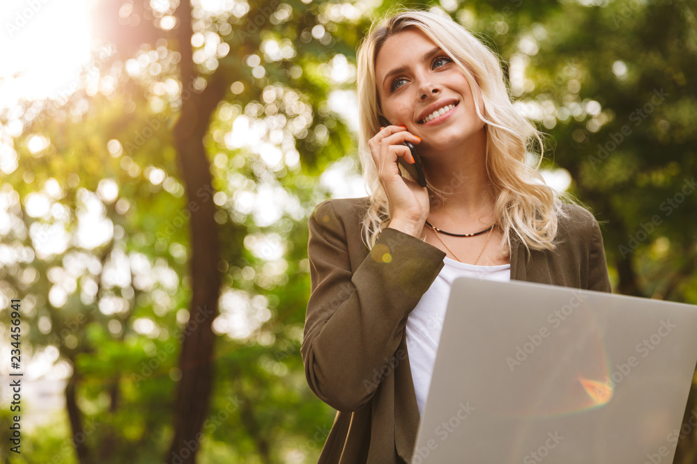 Image of pretty woman 20s using silver laptop and talking on mobile phone, while sitting on bench in park Image of pretty woman 20s using silver laptop and talking on mobile phone, while sitting on bench in park