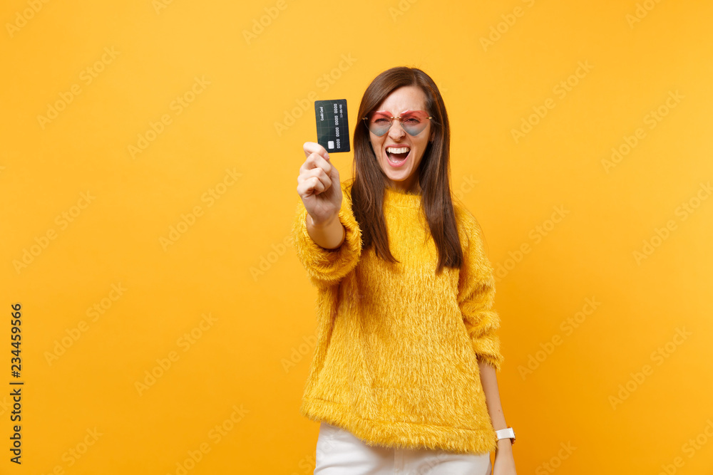 Portrait of funny crazy young woman in fur sweater and heart eyeglasses showing credit card on camera isolated on bright yellow background. People sincere emotions lifestyle concept. Advertising area.