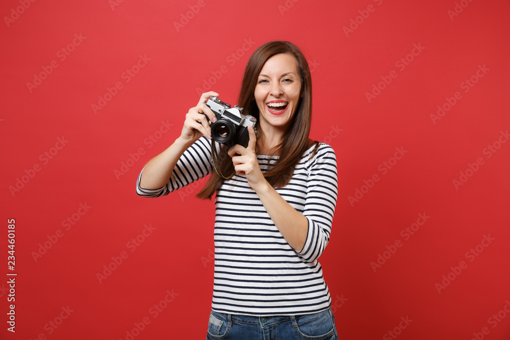 Obraz premium Portrait of joyful beautiful young woman in striped clothes holding retro vintage photo camera isolated on bright red wall background. People sincere emotions, lifestyle concept. Mock up copy space.