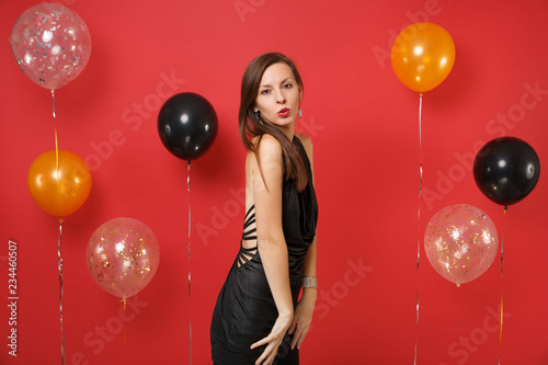 Passionate young woman in little black dress celebrating blowing lips on bright red background air balloons. Valentine's International Women's Day Happy New Year birthday mockup holiday party concept.