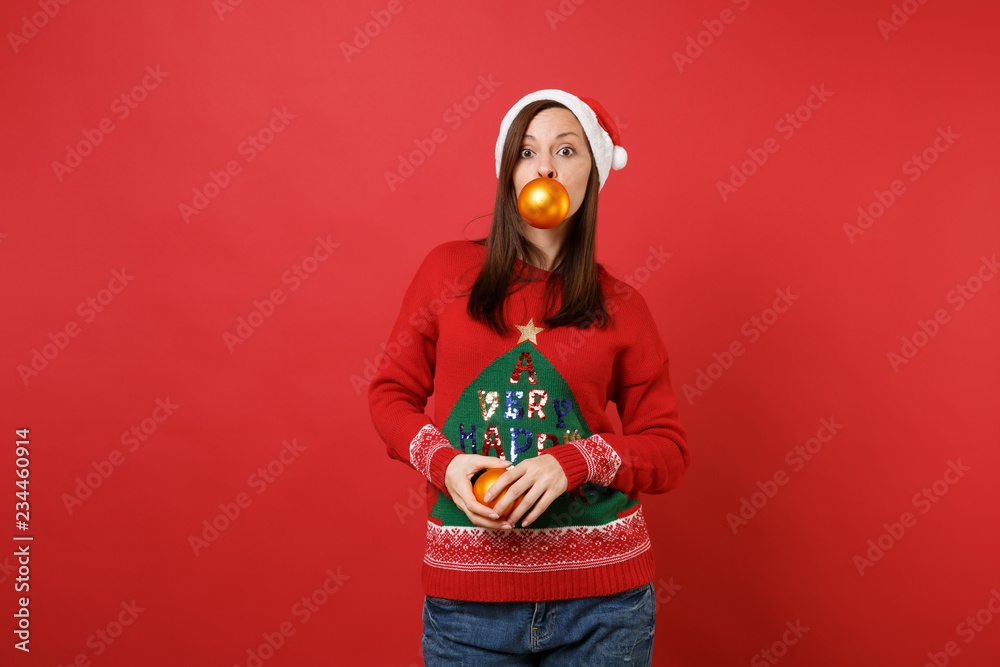 Attractive young Santa girl in sweater, Christmas hat holding yellow tree toys in mouth and hands isolated on red background. Happy New Year 2019 celebration holiday party concept. Mock up copy space.