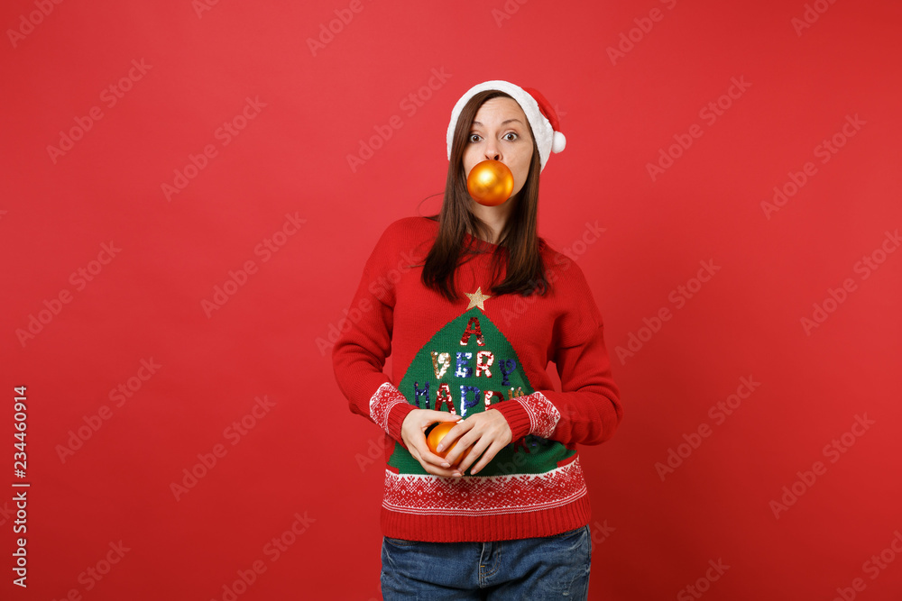 Fototapeta premium Attractive young Santa girl in sweater, Christmas hat holding yellow tree toys in mouth and hands isolated on red background. Happy New Year 2019 celebration holiday party concept. Mock up copy space.