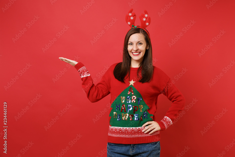 Pretty young Santa girl in knitted sweater, fun decorative deer horns on head pointing hand aside isolated on red background. Happy New Year 2019 celebration holiday party concept. Mock up copy space. Pretty young Santa girl in knitted sweater, fun decorative deer horns on head pointing hand aside isolated on red background. Happy New Year 2019 celebration holiday party concept. Mock up copy space.