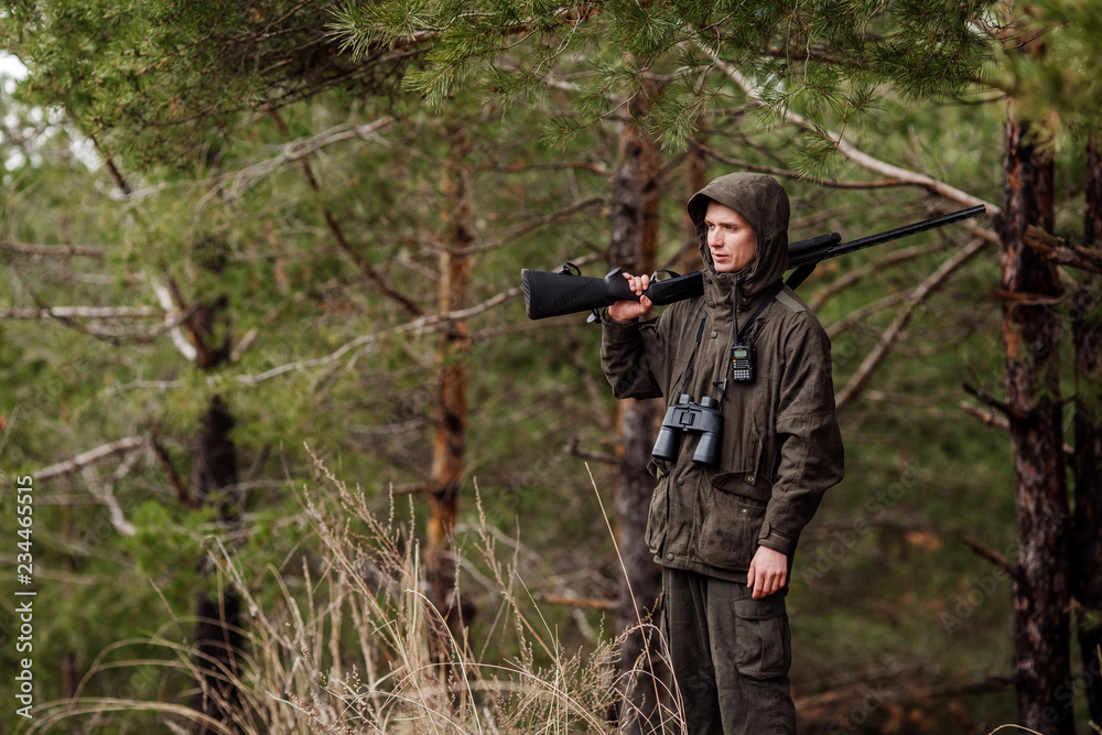 Naklejka premium male hunter with binoculars ready to hunt, holding gun and walking in forest.