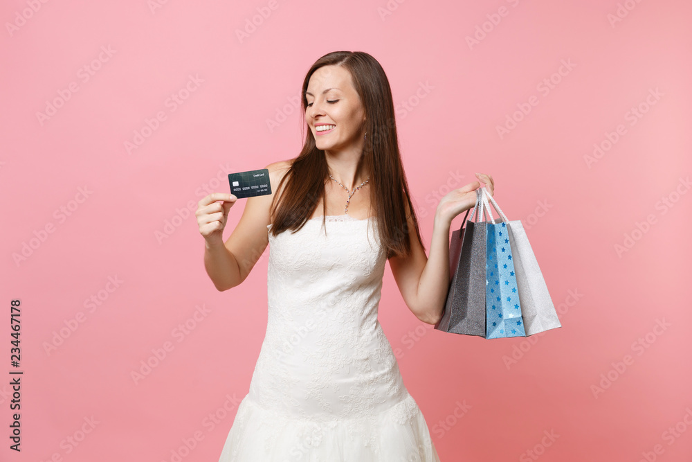 Portrait of tender bride woman in wedding dress holding credit card, multi colored packages bags with purchases after shopping isolated on pink background. Organization of wedding concept. Copy space. Portrait of tender bride woman in wedding dress holding credit card, multi colored packages bags with purchases after shopping isolated on pink background. Organization of wedding concept. Copy space.
