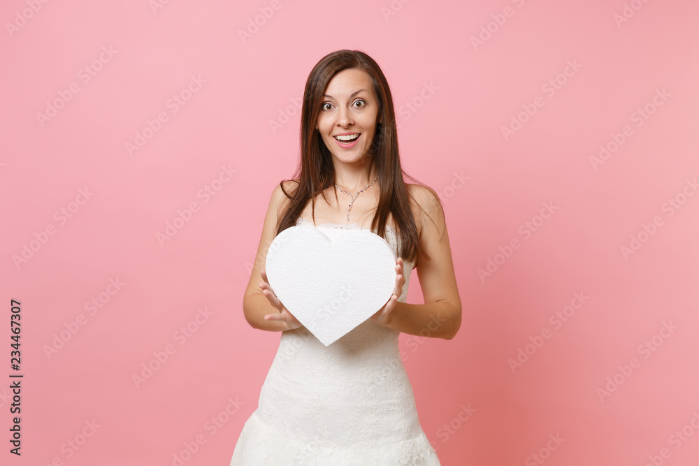 Portrait of smiling beautiful bride woman in elegant white wedding dress standing holding white heart with copy space isolated on pastel pink background. Wedding celebration concept. Advertising area. Portrait of smiling beautiful bride woman in elegant white wedding dress standing holding white heart with copy space isolated on pastel pink background. Wedding celebration concept. Advertising area.