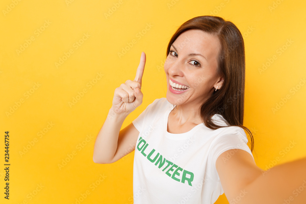 Selfie image of happy smiling satisfied woman in white t-shirt with written inscription green title volunteer isolated on yellow background. Voluntary free assistance help, charity grace work concept. Selfie image of happy smiling satisfied woman in white t-shirt with written inscription green title volunteer isolated on yellow background. Voluntary free assistance help, charity grace work concept.