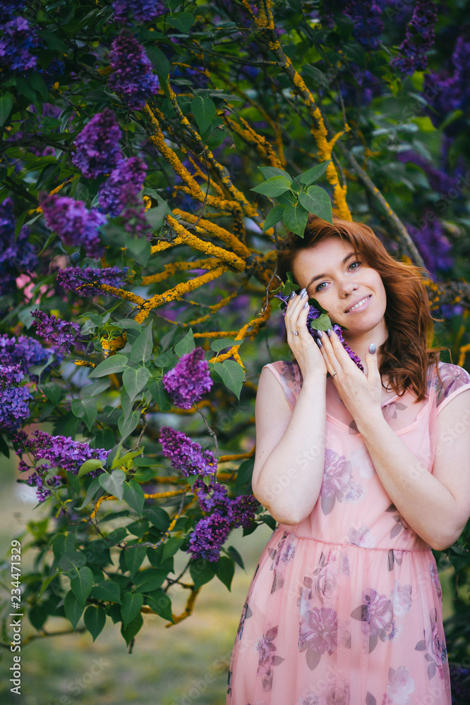 Pretty young brunette girl posing in blooming lilac tree with dark blue flowers outdoor in park. Nice looking tender happy cheerful woman wearing pink stylish feminine dress. Cute female adore nature. Pretty young brunette girl posing in blooming lilac tree with dark blue flowers outdoor in park. Nice looking tender happy cheerful woman wearing pink stylish feminine dress. Cute female adore nature.