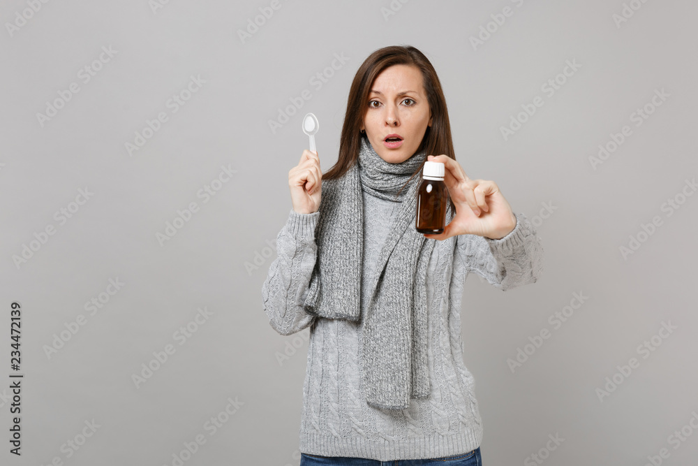 Shocked young woman in gray sweater, scarf holding pour liquid medicine, spoon isolated on grey wall background. Healthy lifestyle ill sick disease treatment, cold season concept. Mock up copy space. Shocked young woman in gray sweater, scarf holding pour liquid medicine, spoon isolated on grey wall background. Healthy lifestyle ill sick disease treatment, cold season concept. Mock up copy space.