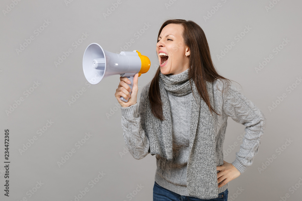 Crazy young woman in gray sweater, scarf scream in megaphone isolated on grey background, studio portrait. Healthy fashion lifestyle, people sincere emotions, cold season concept. Mock up copy space. Crazy young woman in gray sweater, scarf scream in megaphone isolated on grey background, studio portrait. Healthy fashion lifestyle, people sincere emotions, cold season concept. Mock up copy space.