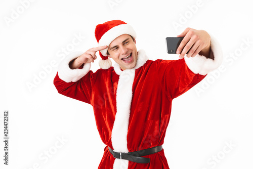 Image of optimistic man 30s in santa claus costume holding smartphone and taking selfie, isolated on white background in studio