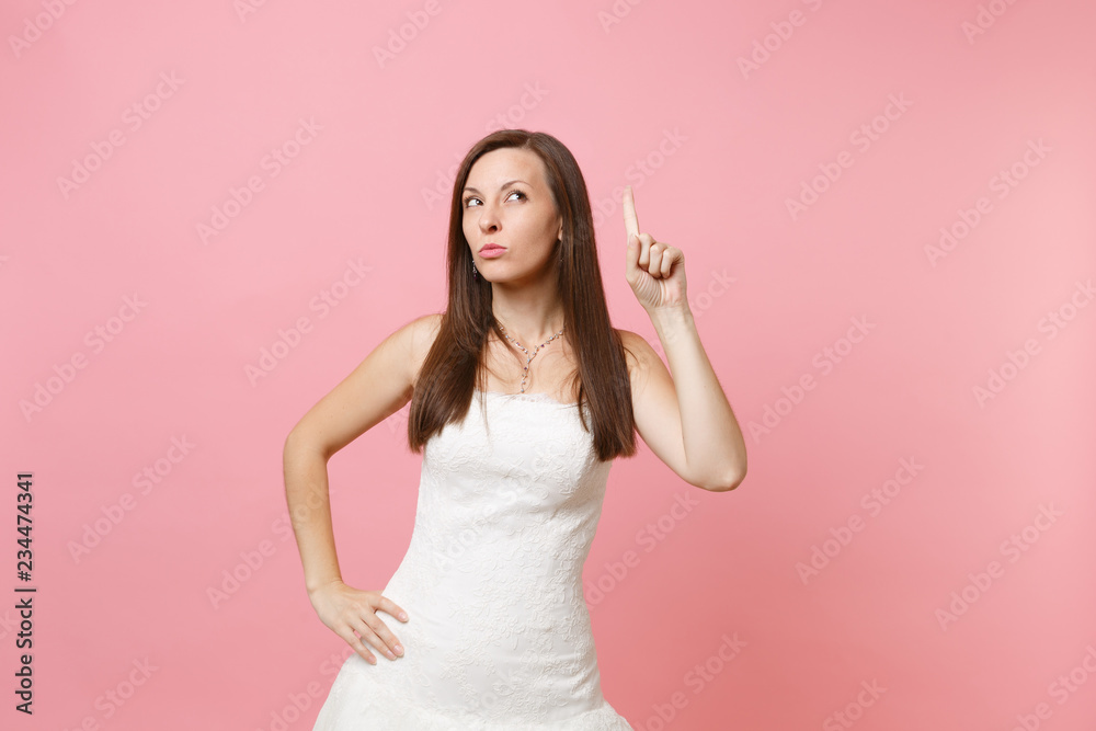 Portrait of concerned strict bride woman in elegant white wedding dress standing pointing index finger up isolated on pastel pink background. Wedding celebration concept. Copy space for advertisement. Portrait of concerned strict bride woman in elegant white wedding dress standing pointing index finger up isolated on pastel pink background. Wedding celebration concept. Copy space for advertisement.